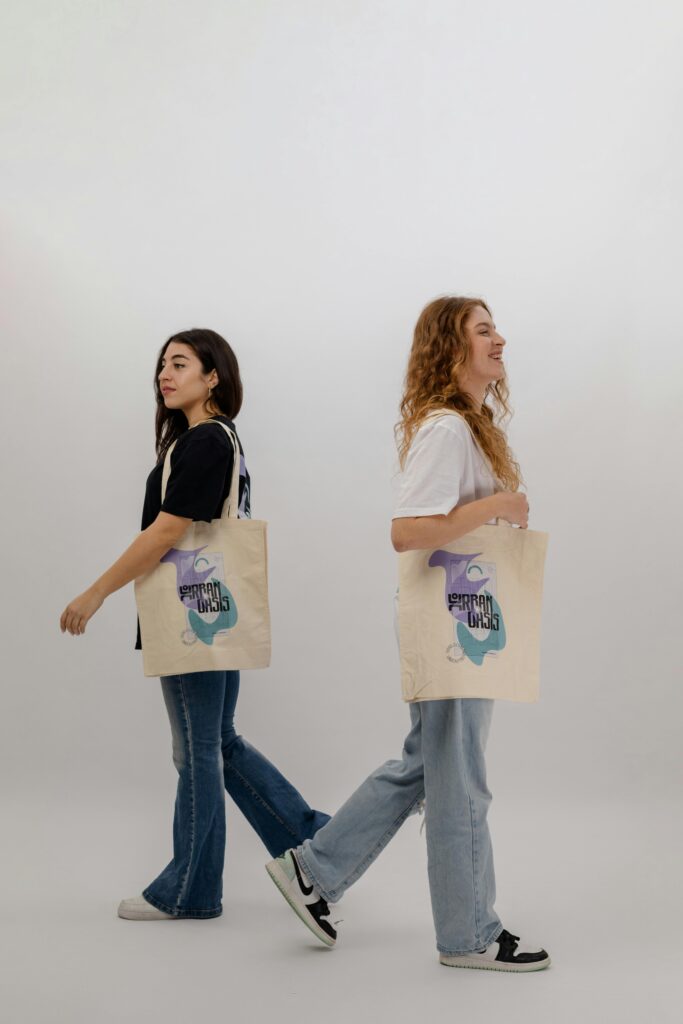 Two young women in jeans showcase graphic tote bags in a studio setting, emphasizing casual fashion.